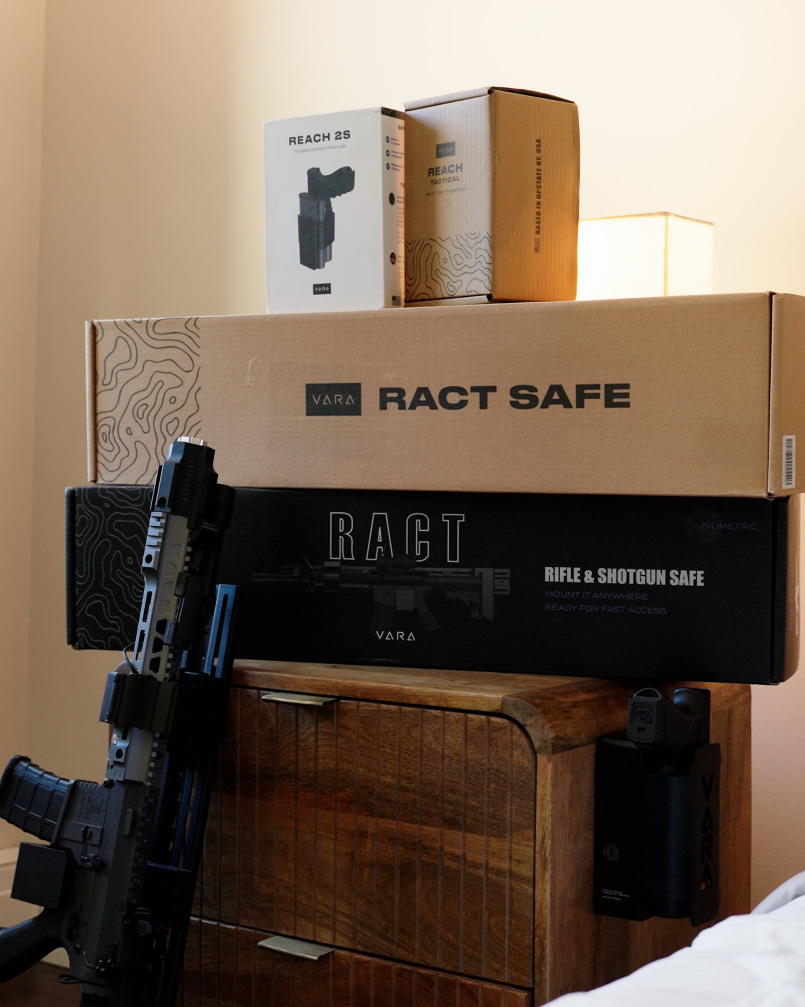 RACT Safe, RACT Biometric, Reach Tactical and Reach 2S boxes stacked on a nightstand, a Reach safe mounted on its side and an AR leaning against it