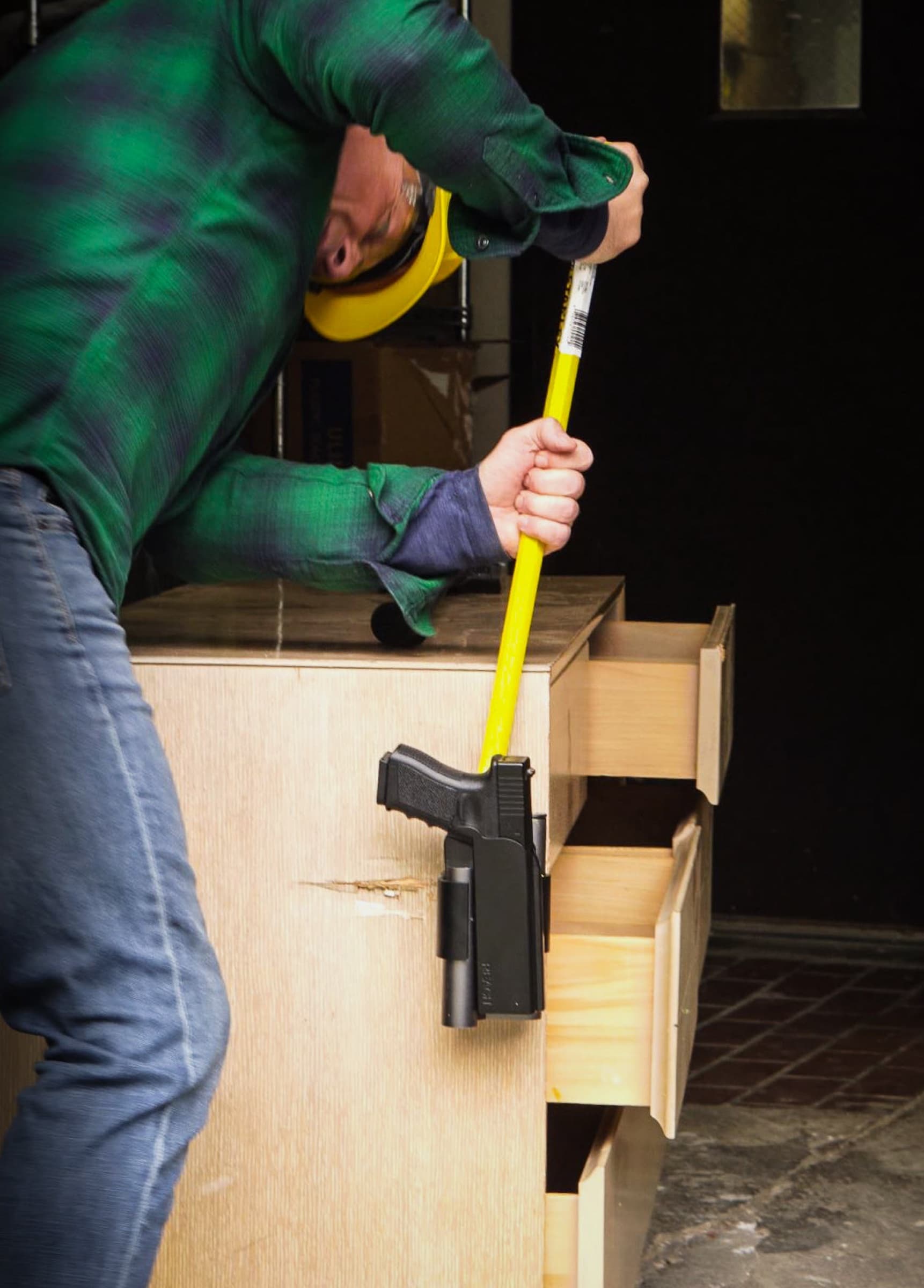 A man prying at a Reach 2S mounted on a dresser with a long pry bar, the pistol still locked in place