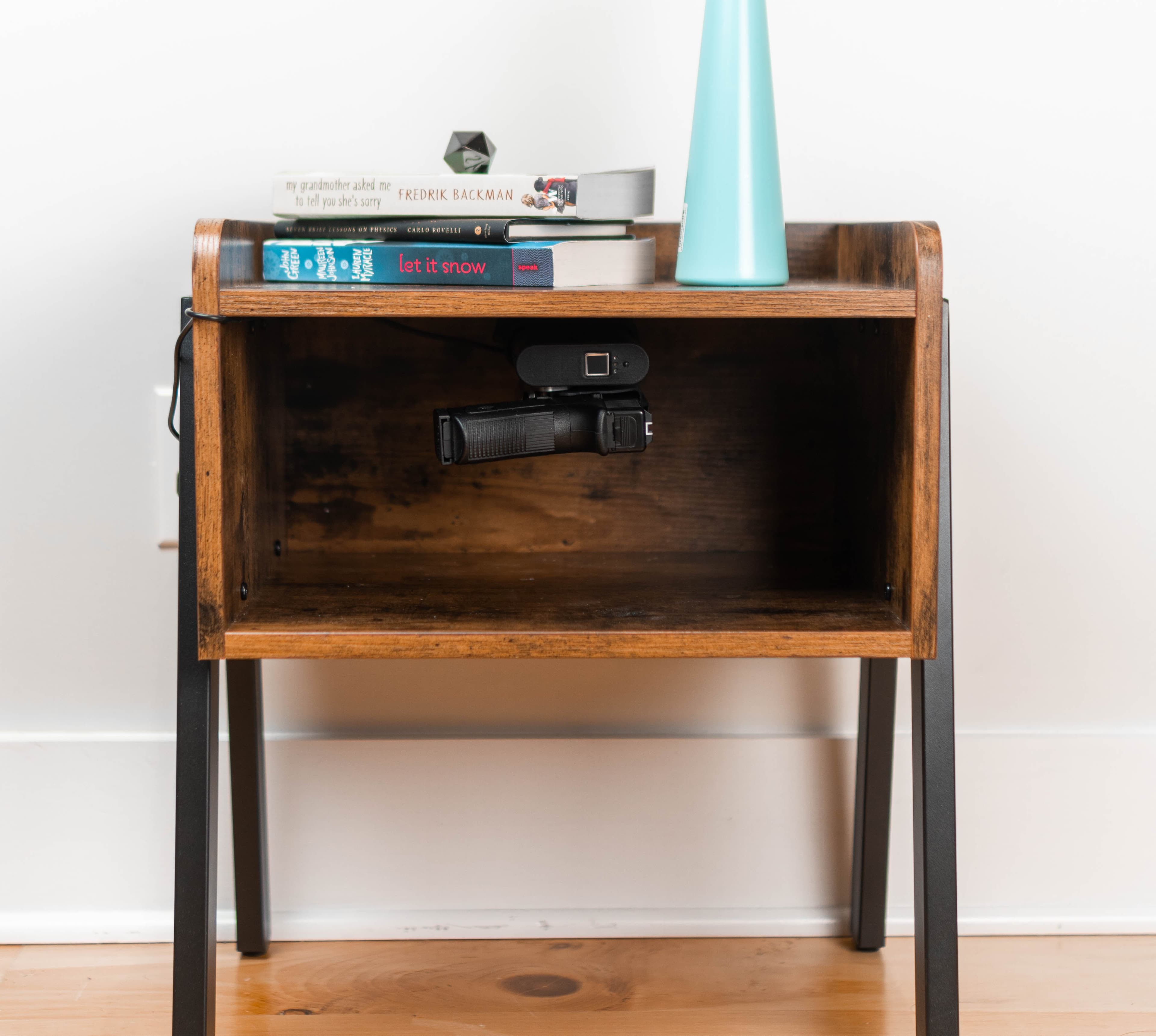 A Reach safe mounted under the shelf of a wooden nightstand, holding a handgun, with books and a lamp on top