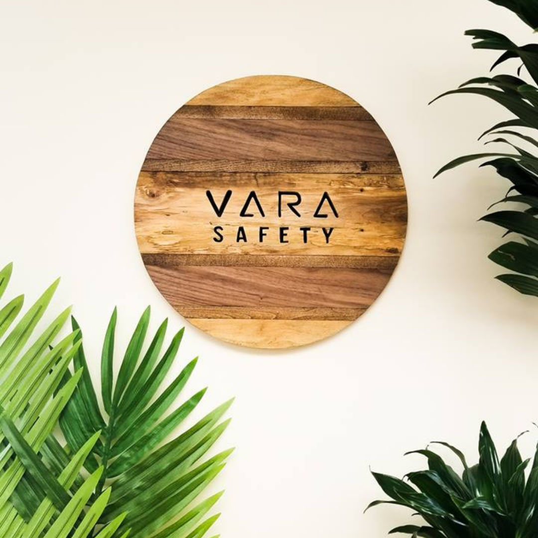 A wooden glazed sign with the Vara Safety named etched into the surface. There are bright green plants surrounding the sign.